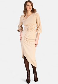 Abito avvolgente beige con lunghe maniche puff, dettaglio arricciato sui lati, orlo asimmetrico e abbinato a stivali con tacco marrone scuro.