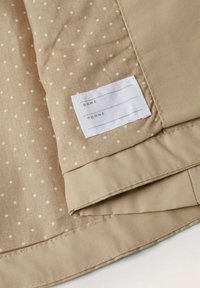 Beige tyg med vita prickar, med en sytt etikett för 'NAMN' och 'TELEFON.' Slät textur med snygg sömnad.