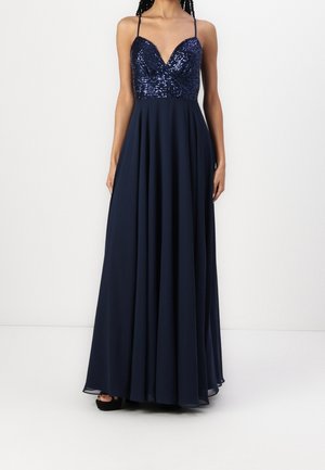 Nainen pukeutuneena tummansiniseen mekkoon, jossa paljettikoristeinen yläosa, spagettiolkaimet, syvä V-pääntie ja laskeutuva lattiaan ulottuva helma, seisoo valkoista taustaa vasten.