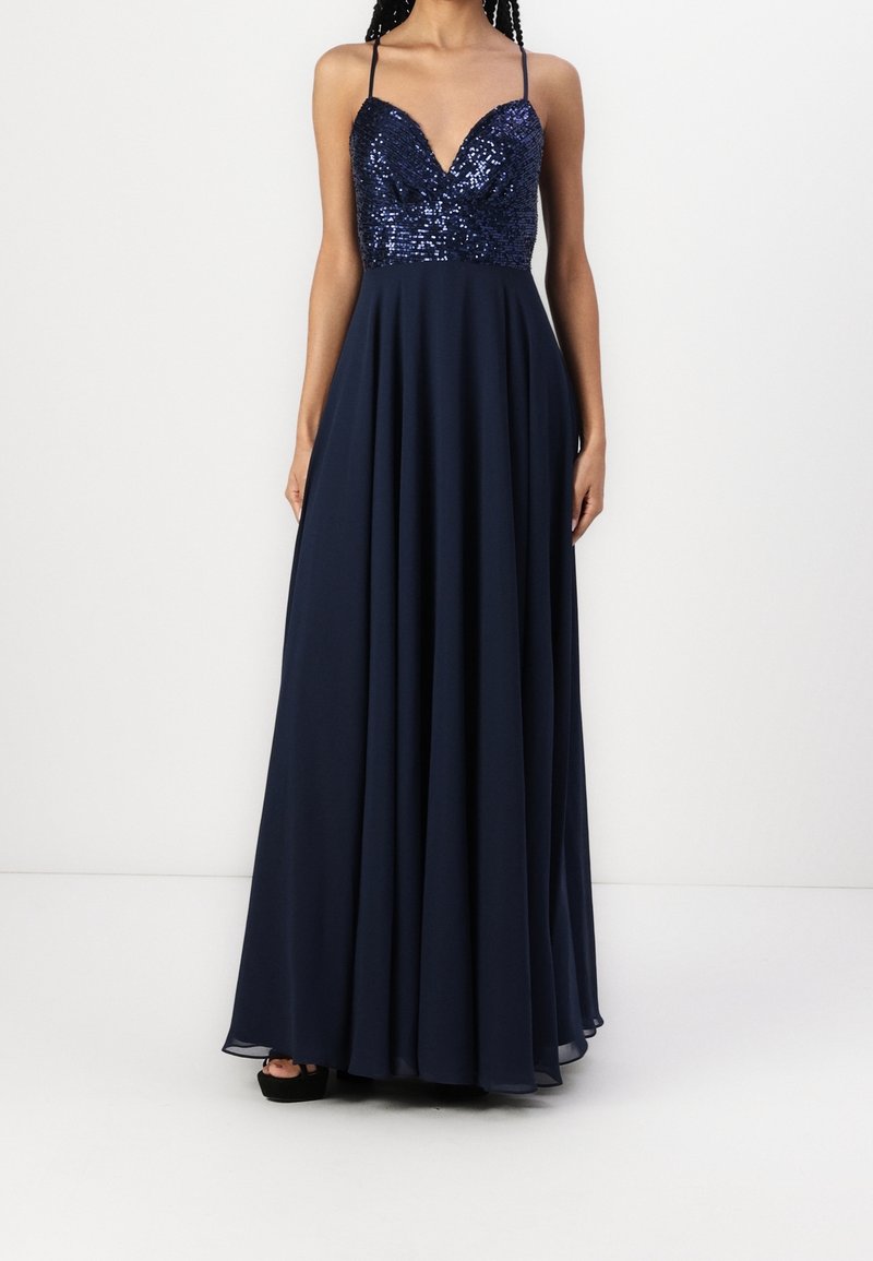 Femme portant une robe bleu marine avec un corsage à sequins, des bretelles spaghetti, un profond décolleté en V et une jupe longue fluide, se tenant devant un fond blanc.