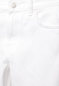 Gros plan sur un jean en denim blanc montrant le tissu texturé, la poche avant avec rivet, passants de ceinture et fermeture par bouton en métal.