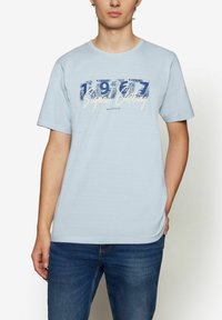 Šviesiai mėlynas trumpas rankovės marškinėliai su apykakle. Priekyje yra grafika su tamsiai mėlyna ir kreminės spalvos tekstu „1977 Signal Clothing“. 
