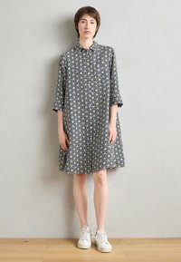 Vestito a righe in bianco e nero con maniche a tre quarti, chiusura con bottoni, vestibilità comoda e sneakers bianche su un pavimento di legno.
