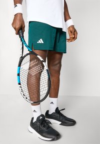 Shorts de tennis texturés verts avec logo Adidas blanc, chaussures de sport noires, chaussettes blanches et une raquette de tennis noire et bleue.