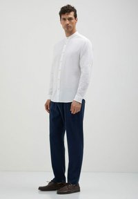 Camicia bianca in lino con colletto alla cinese, abbinata a pantaloni blu scuro e mocassini marroni. Design semplice e pulito.