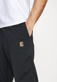 Pantalones deportivos negros con bolsillos laterales, con un parche de logo de Nike multicolor. La tela es suave y tiene un ligero brillo.