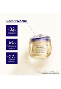 Shiseido Vital Perfection-crème i en rund guldfärgad burk med ett blått lock med gradient. Produktnamn och effektivitetsegenskaper framhävs i blå rutor.