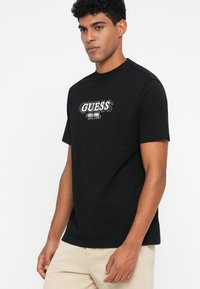 Camiseta de algodón negra de mangas cortas, con cuello redondo, que presenta un estampado gráfico blanco que dice "GUESS" junto con las fechas de establecimiento.