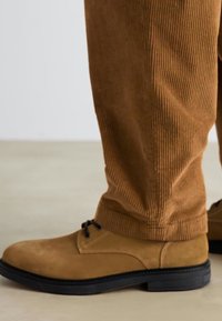 Chaussures en daim marron avec des semelles en caoutchouc noires, dotées de lacets ronds et de détails de couture. Assorties à un pantalon en velours côtelé marron.