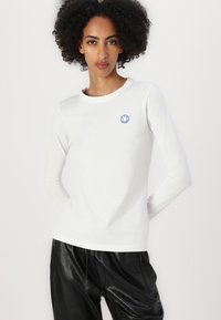 Camicia bianca a maniche lunghe in cotone, con scollatura rotonda e un piccolo logo di un sorriso blu sul petto. Abbinata a pantaloni in pelle nera.