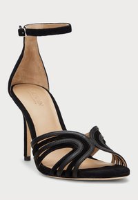 Μαύρη σουέτ ψηλοτάκουνη σαγιονάρα με λουράκι στον αστράγαλο. Διαθέτει καμπύλο σχέδιο λουριού και ανοιχτό δάχτυλο. Λείος μπεζ πάτος και λεπτός τακούνι στιλέτο.