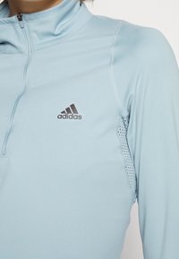 Helles blaues sportliches Halb-Zip-Oberteil mit hohem Kragen, das atmungsaktive Netzbesätze und ein dezentes schwarzes Adidas-Logo auf der Brust aufweist.