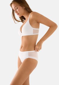 Mujer joven ajustándose un sujetador blanco y ropa interior a juego con paneles laterales de malla, de pie de perfil contra un fondo claro y liso.