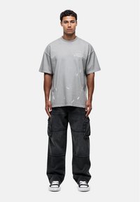 T-shirt gris oversize avec des détails usés, associé à un pantalon cargo noir avec des poches latérales et une coupe décontractée. Des baskets noires complètent le look.
