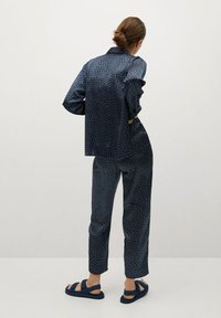 Frau mit einem Dutt, die ein dunkelblaues gemustertes Langarmhemd und passende Hose trägt, mit Händen in den Taschen steht, sichtbare marineblaue Sandalen.
