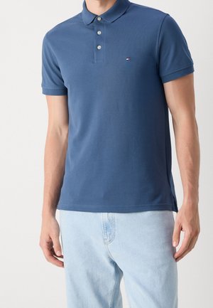 Homme portant un polo bleu à manches courtes avec des boutons et un jean bleu clair, debout devant un fond uni.
