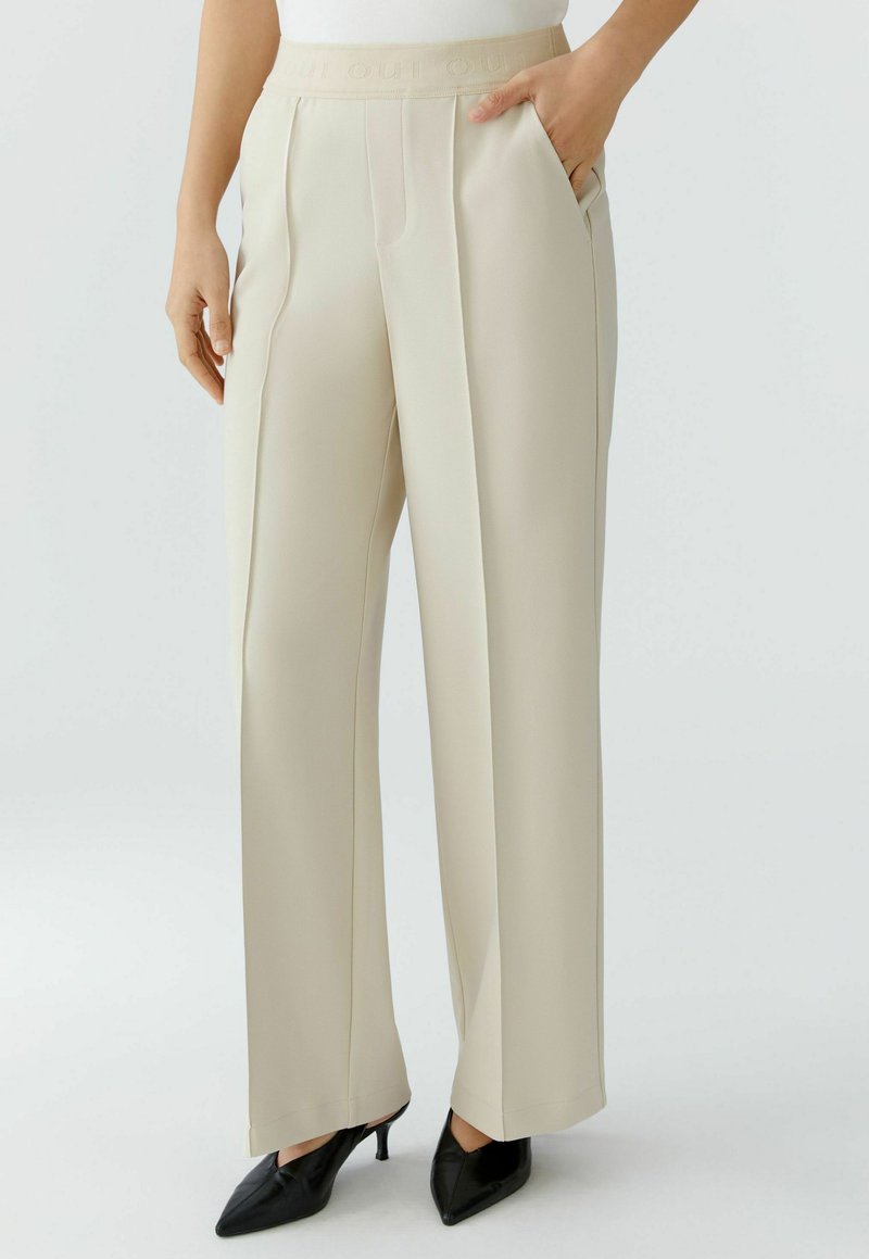 Beige tailored trousers with a high waistband, front pleats, side pockets, and a smooth texture, paired with black pointed heels.