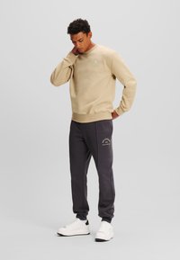 Sudadera beige con cuello redondo, puños acanalados y detalle de logo; combinada con joggers gris oscuro que presentan texto bordado; zapatillas blancas.