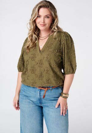 Femme souriante aux cheveux blonds ondulés portant un chemisier floral texturé vert olive, un jean bleu, et des bijoux et bracelets dorés superposés.