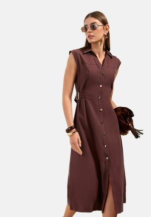 Naine, kes kannab varrukateta pruunikat nupuga keskmise pikkusega kleiti, päikeseprille, käevõrusid ja hoiab pruuni tutiga õlakotti.