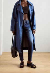 Trench de mezclilla en azul profundo, corte holgado con cuello ancho y cierres de botones, combinado con jeans oscuros y sandalias negras de tacón bloque.