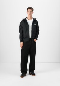 Svart vindjakke med glidelås-lommer, brukt over en hvit skjorte, kombinert med svarte vide bukser og mørke sneakers. Studio bakgrunn.
