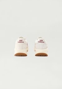 Chaussures de sport blanches avec une tige en daim, arborant un logo bordeaux, une semelle intermédiaire beige amortie et une semelle extérieure en caoutchouc avec motifs antidérapants.