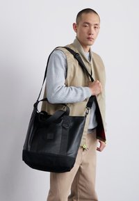 Sac fourre-tout en cuir noir avec finition texturée, doté de poignées doubles, d'une sangle réglable et d'un petit logo sur le devant.