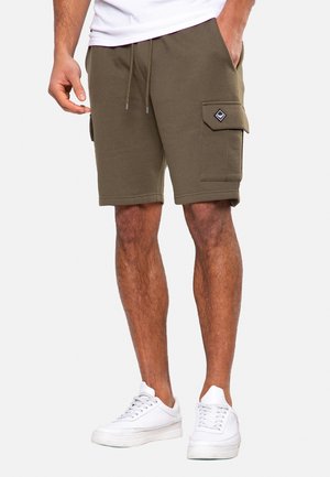 Hombre usando pantalones cortos cargo verde oliva con bolsillos laterales y zapatillas blancas, de pie con una mano en el bolsillo.