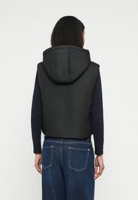 Μαύρο καπιτονέ γιλέκο με κουκούλα, με κομψό σχέδιο και απαλή υφή. Φορεμένο πάνω από μ�αύρο μπλουζάκι με μακριά μανίκια και μπλε τζιν.