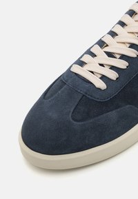 Suede navy sneaker with a rounded toe, cream laces, and a beige rubber sole. Features a smooth texture and minimalistic design elements.