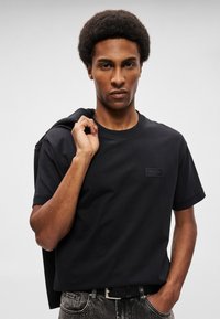 Μαύρο μπλουζάκι φτιαγμένο από βαμβάκι, με στρογγυλή λαιμόκοψη και κοντά μανίκια, που έχει ένα ορθογώνιο λογότυπο στο στήθος, σε συνδυασμό με τζιν.