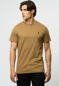 Tricou din bumbac bej, cu un design ajustat, guler rotund și logo negru pe piept. Asortat cu blugi negri, evidențiind un look casual.