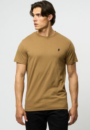 Tricou din bumbac bej, cu un design ajustat, guler rotund și logo negru pe piept. Asortat cu blugi negri, evidențiind un look casual.