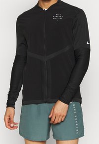 Man in een zwart Nike Running Division longsleeve zipjack en groene geperforeerde Nike Running shorts, met ontspannen handen naast zijn lichaam.