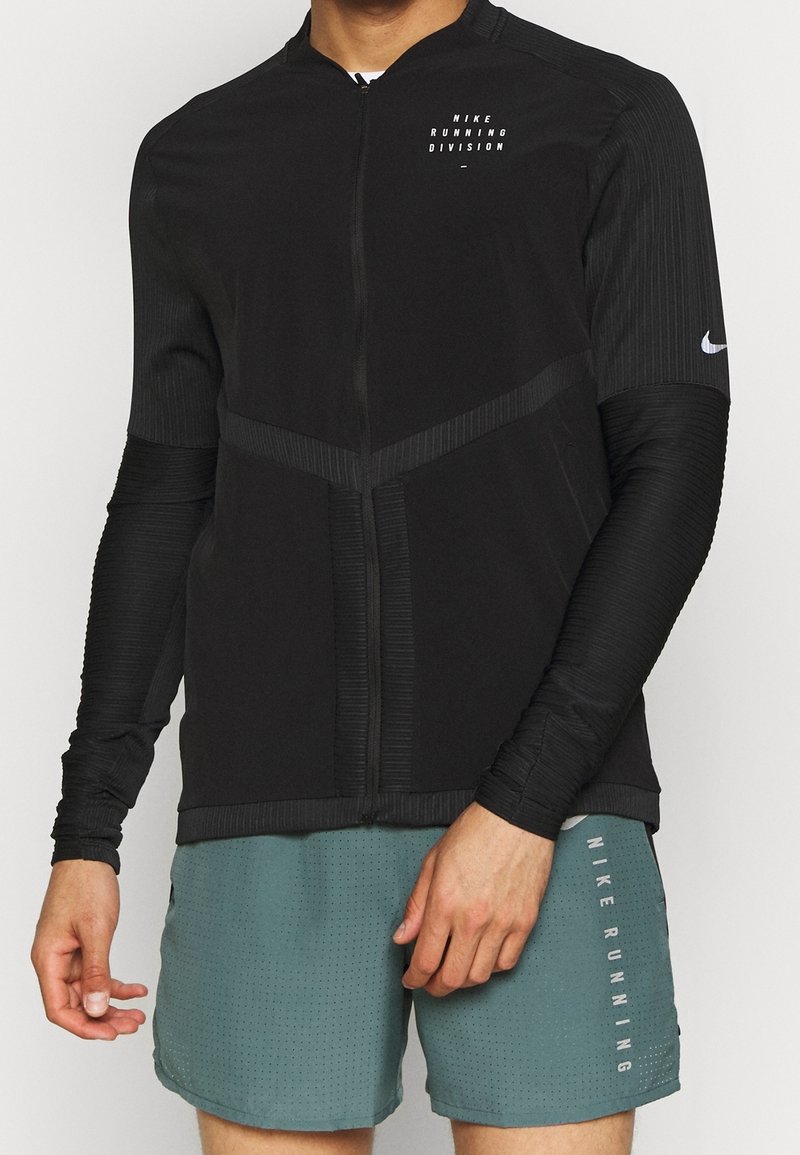 Man in een zwart Nike Running Division longsleeve zipjack en groene geperforeerde Nike Running shorts, met ontspannen handen naast zijn lichaam.