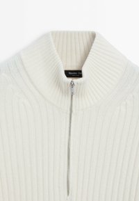 Pull en maille côtelée crème avec col haut et demi-fermeture éclair argentée, montrant une étiquette de marque visible à l'intérieur du col.