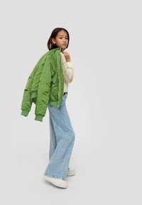 Groene, gevoerde bomberjack met ribgebreide boorden, gedragen over een gebreide crèmekleurige trui. Slim, uitlopende blauwe jeans en witte platform sneakers maken de look compleet.