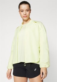 Light yellow, zip-up jacket with elastic cuffs and drawstring hem, worn over a light top and black shorts, featuring a subtle logo.