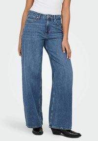 Jean en denim à jambes larges de couleur bleu clair, comprenant cinq poches, une braguette à boutons, et des détails cousus. Le tissu est doux avec une finition légèrement délavée.