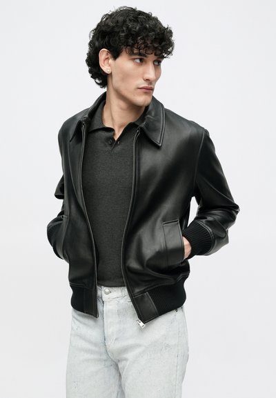 Μαύρο δερμάτινο μπουφάν τύπου bomber με γιακά, ριμπ μανσέτες και τσέπες με φερμουάρ, φορεμένο πάνω από σκούρο γκρι πόλο και ανοιχτόχρωμο τζιν.