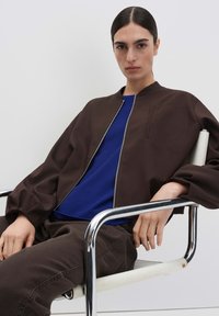 Giacca bomber marrone con zip frontale e dettaglio tasca, indossata sopra una maglia blu. Modello seduto su una sedia cromata, pantaloni scuri visibili.