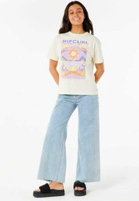 T-shirt à manches courtes couleur crème avec un graphisme coloré de soleil et de vagues, associé à un jean large bleu clair et des sandales à plateforme noires.