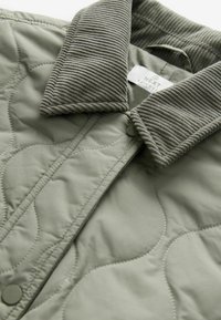Olivgrüne, gesteppte Jacke mit strukturiertem Cordkragen und Druckknöpfen, die das gestickte Muster und das Innenetikett zeigt.