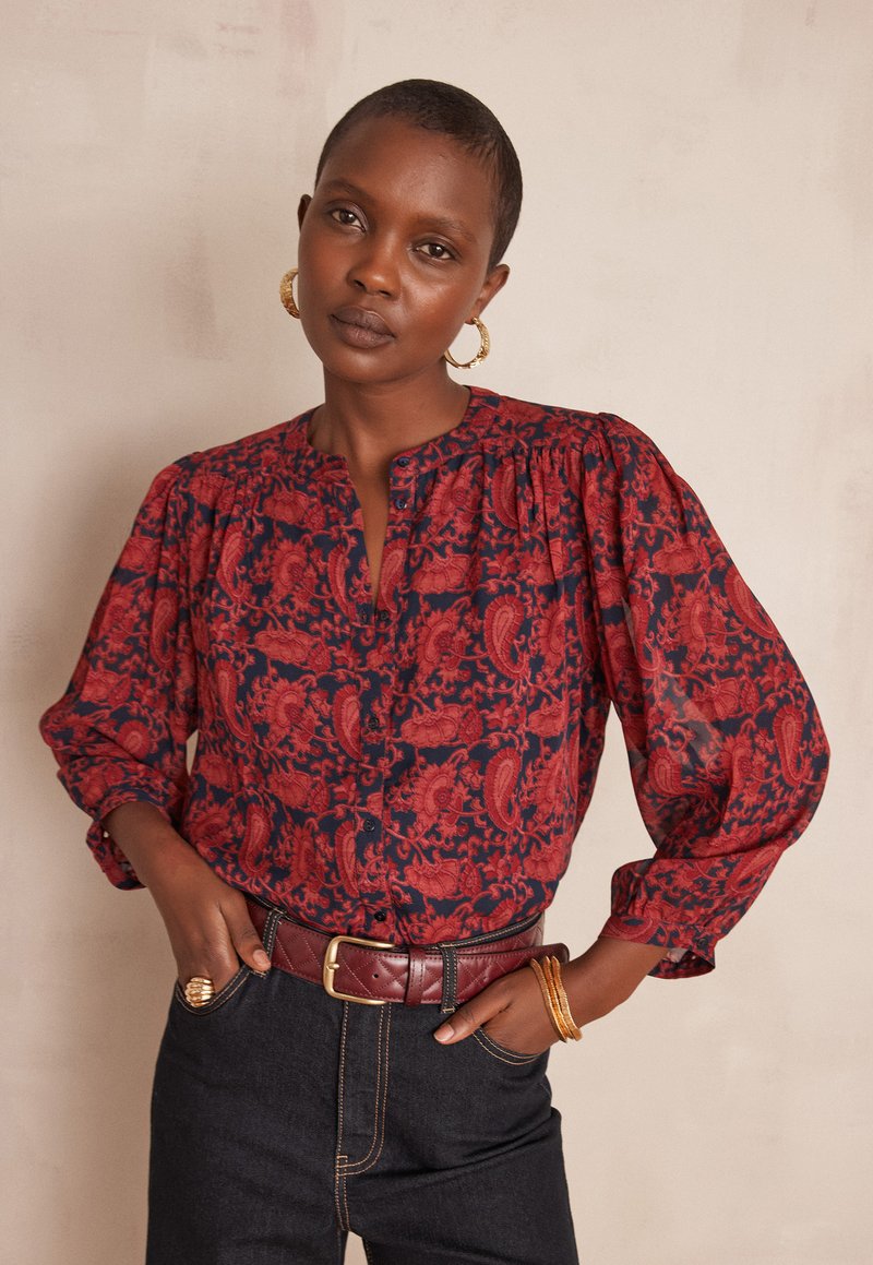 Blusa rossa e blu navy con motivo, con chiusura frontale a bottoni, spalle arricciate e maniche lunghe, indossata con jeans scuri a vita alta e cintura bordeaux.