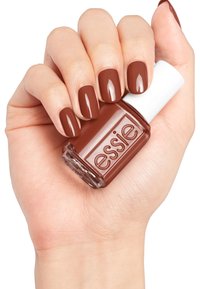 Brauner Nagellack in einer klaren Glasflasche, die in einer Hand gehalten wird. Der Lack hat ein glänzendes Finish und der Markenname "essie" ist auf der Flasche sichtbar.