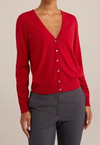 Röd cardigan i mjukt tyg, med djup V-ringning, långa ärmar och silverknappar längs fronten; kombinerad med grå byxor.