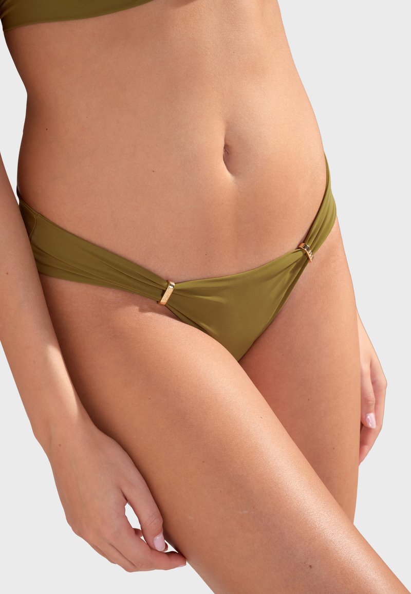 Gros plan sur une personne portant un bas de bikini vert olive avec des accents d'anneaux dorés sur les hanches, sur un fond clair uni.