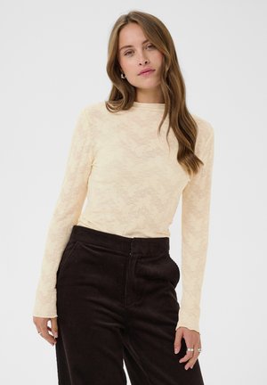 Femme aux cheveux châtain clair porte un haut crème texturé à manches longues et un pantalon en velours côtelé marron foncé taille haute, debout devant un fond uni.