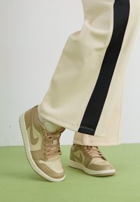 Baskets Nike beiges avec un bout perforé, un col rembourré et des accents marron. Portées avec un pantalon clair présentant une bande latérale noire.